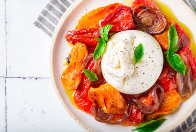 Burrata cheese with baked tomatoes, pepper, red onion and fresh basil on white tile background. 