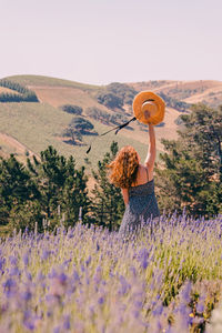 Woman standing in a purple lavender field, holding straw hat with ribbons, view from the back