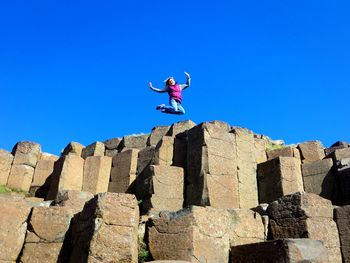 Low angle view of woman jumping on rocks against clear blue sky