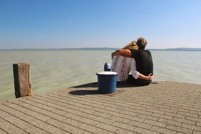 Rear view of couple sitting with arms around on pier by sea