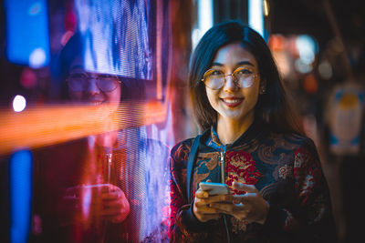 Portrait of smiling young woman using smart phone by window with reflection at night 