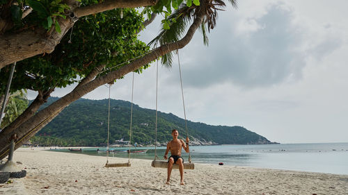 Shirtless man sitting on swing at beach against sky