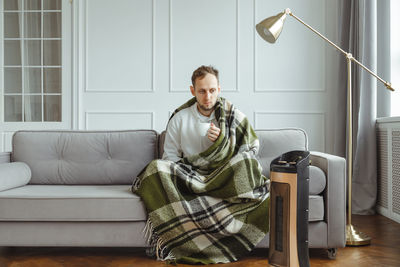 Young man in wrapped in plaid in warm clothes with cup of tea at home. concept of heating season