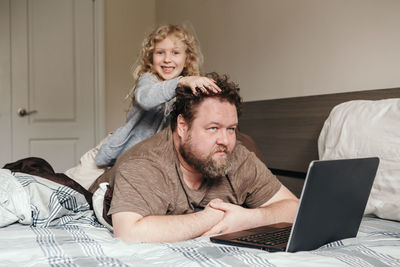 Portrait of daughter sitting on father lying on bed using laptop at home