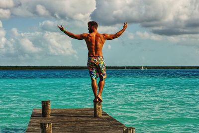 Rear view of shirtless man posing while standing on pier against sky