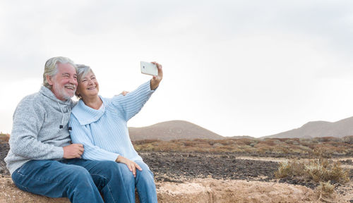 Smiling senior couple taking selfie while sitting at remote location against clear sky