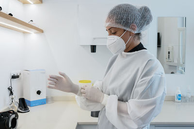 Side view of young female medical worker in white uniform and mask putting on latex gloves while preparing for treatment in modern clinic