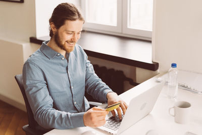 Happy young man with credit card in hand shopping online on laptop, top view