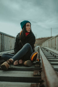 Young woman sitting on railroad track against sky