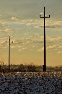 Electricity pylons on field against cloudy sky during sunset