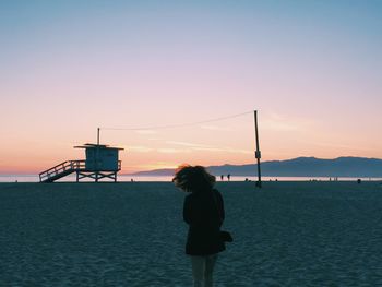 Rear view of woman standing at beach against sky during sunset