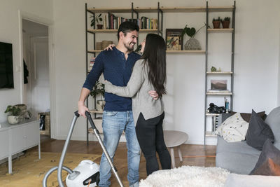 Man embracing woman while using vacuum cleaner in living room