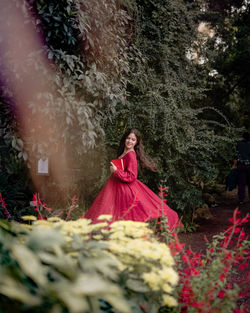 Rare view of a beautiful young woman dancing in a long red dress with book around flowers and trees