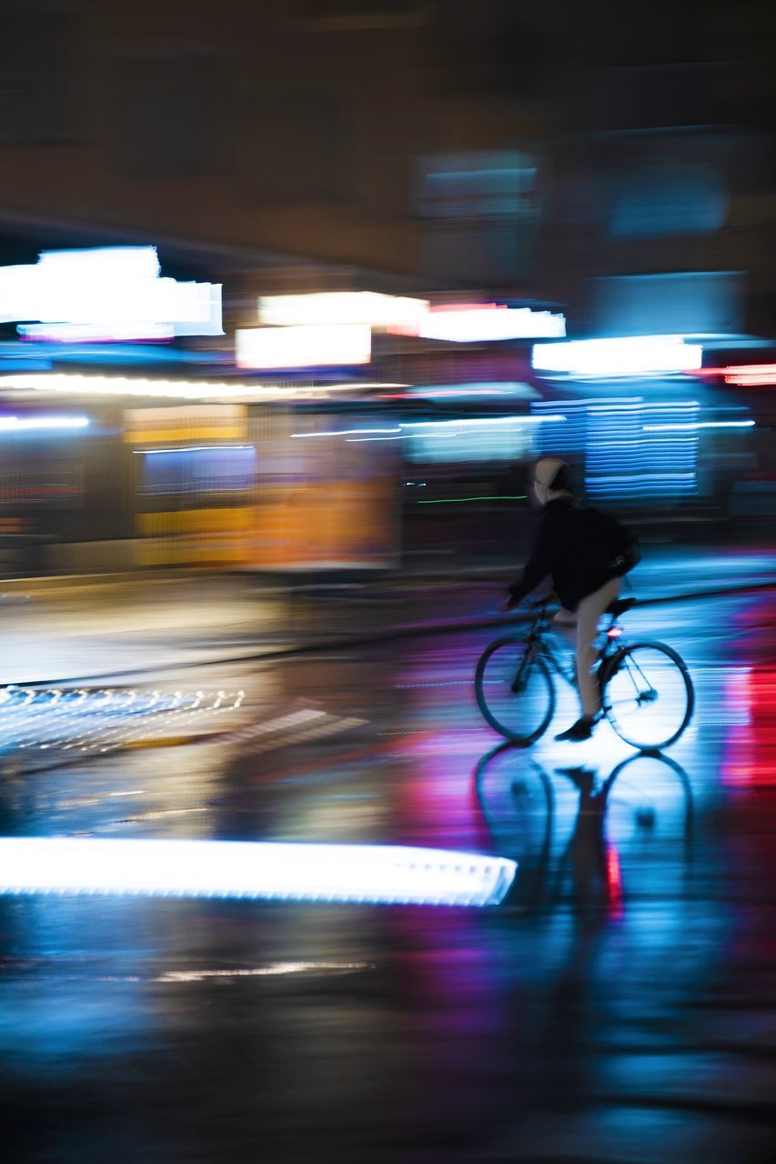 BLURRED MOTION OF MAN RIDING BICYCLE ON ILLUMINATED STREET