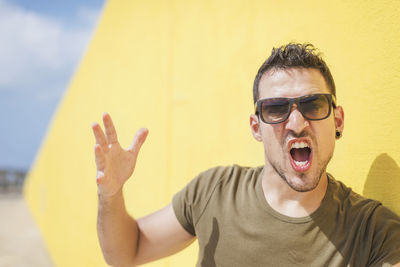 Portrait of man wearing sunglasses shouting while sanding against wall outdoors