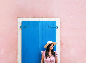 Woman in pink dress standing by blue door, window on pink wall.