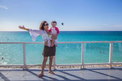 Woman carrying toddler daughter on promenade with sea in background against blue sky