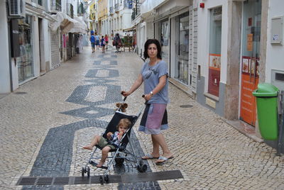 Woman with son sitting in baby stroller on footpath amidst buildings