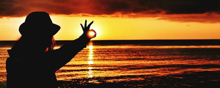 Silhouette woman gesturing ok sign at beach against sky during sunset