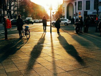 People standing with shadow on footpath during sunset in city