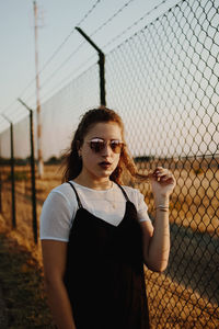 Portrait of young woman standing by fence against sky