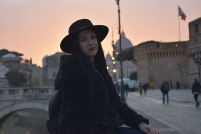 Portrait of young woman standing in city against sky during sunset
