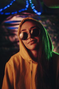 Portrait of smiling young woman wearing sunglasses and hood at illuminated restaurant