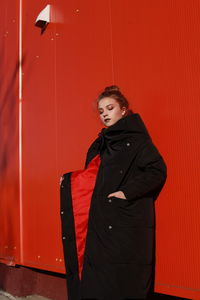 Fashionable teenage girl wearing black warm clothing standing against red wall during sunny day