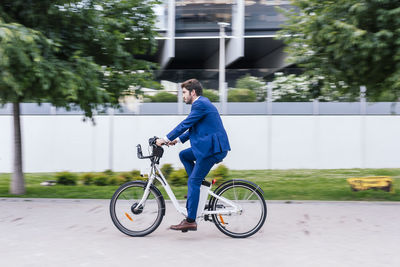 Young smiling man in elegant suit riding electric bicycle on pavement while looking away