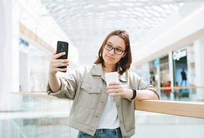 Young teenager girl student in glasses using mobile phone taking selfie at shopping mall