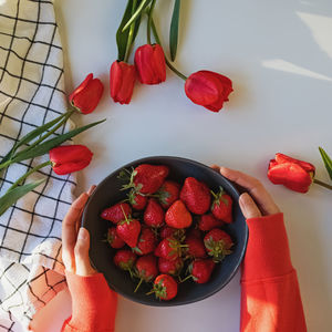Woman's hands holding a bowl of fresh strawberries on the table with red tulips, spring composition