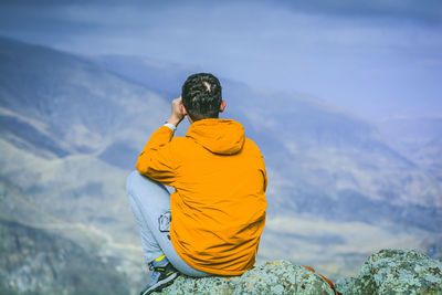 Rear view of man sitting on cliff against mountains