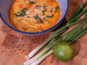 Asian coconut soup with red curry paste in a blue bowl on a wooden block with spring onions, lime.