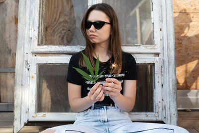 Young woman holding cannabis and illuminated sparkler while sitting against window