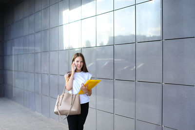Young businesswoman talking on mobile phone while standing by wall