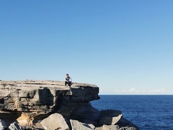 Man sitting on rock formation by sea against blue sky