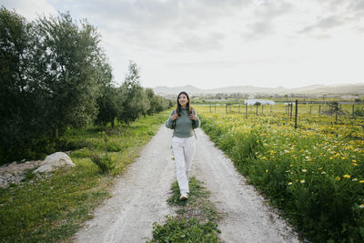 Full length of smiling woman with backpack walking on road by field against sky