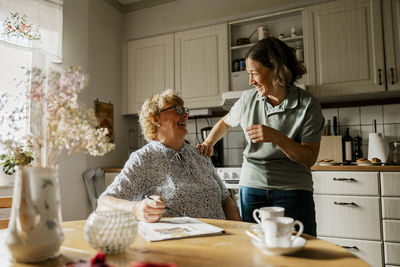Happy female caregiver with elderly woman sitting near dining table in kitchen at home