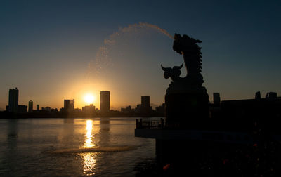 Silhouette carp dragon statue fountain by lake in city against sky during sunset