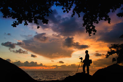 Silhouette man with camera on rock by sea against sky during sunset
