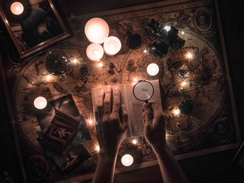 Cropped hands reading book with magnifying glass at illuminated table