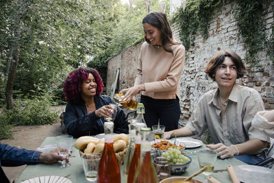 Woman serving drink to female friend sitting at dining table during dinner party in back yard