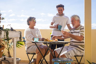 Senior couple with grandson eating food at table on building terrace against sky