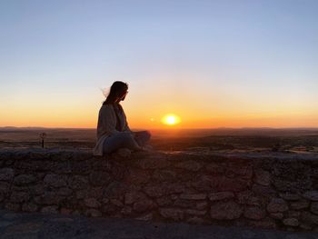 Woman sitting on stone wall against sky during sunrise