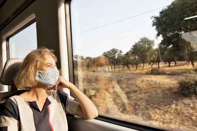 Pensive female traveler wearing protective mask sitting on comfortable passenger seat and looking out of window of train