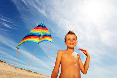 Shirtless boy at beach holding kite against sky