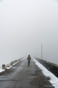 Rear view of person walking on road across a dam. foggy, moody, cold weather.