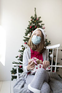 Girl wearing mask holding gifts against christmas tree at home