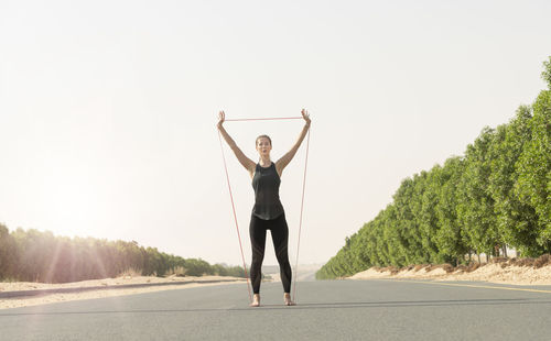 Full length of woman exercising with rubber band on road against clear sky