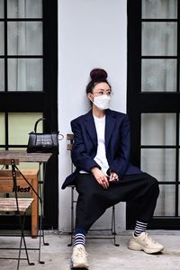 Full length of woman wearing mask looking away while siting by wall